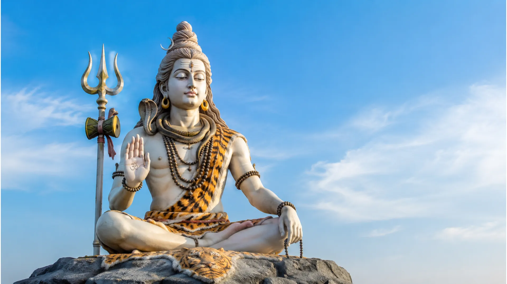 : Large statue of Lord Shiva seated calmly with a trident beside him against a clear blue sky.