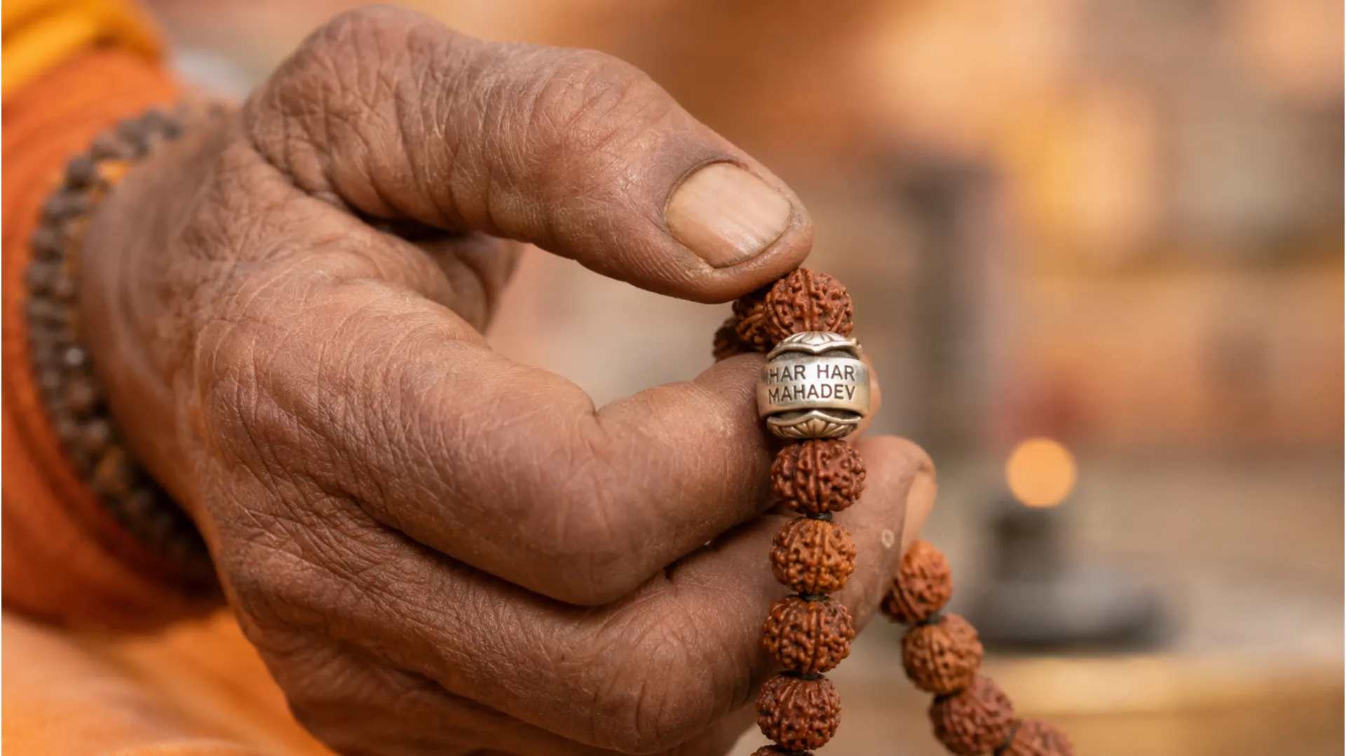 A detailed, macro clear photo camera image of an elderly Sadhu's weathered hand holding a 108-bead Rudraksha mala, with one silver-capped bead clearly engraved with 'HAR HAR MAHADEV'.
