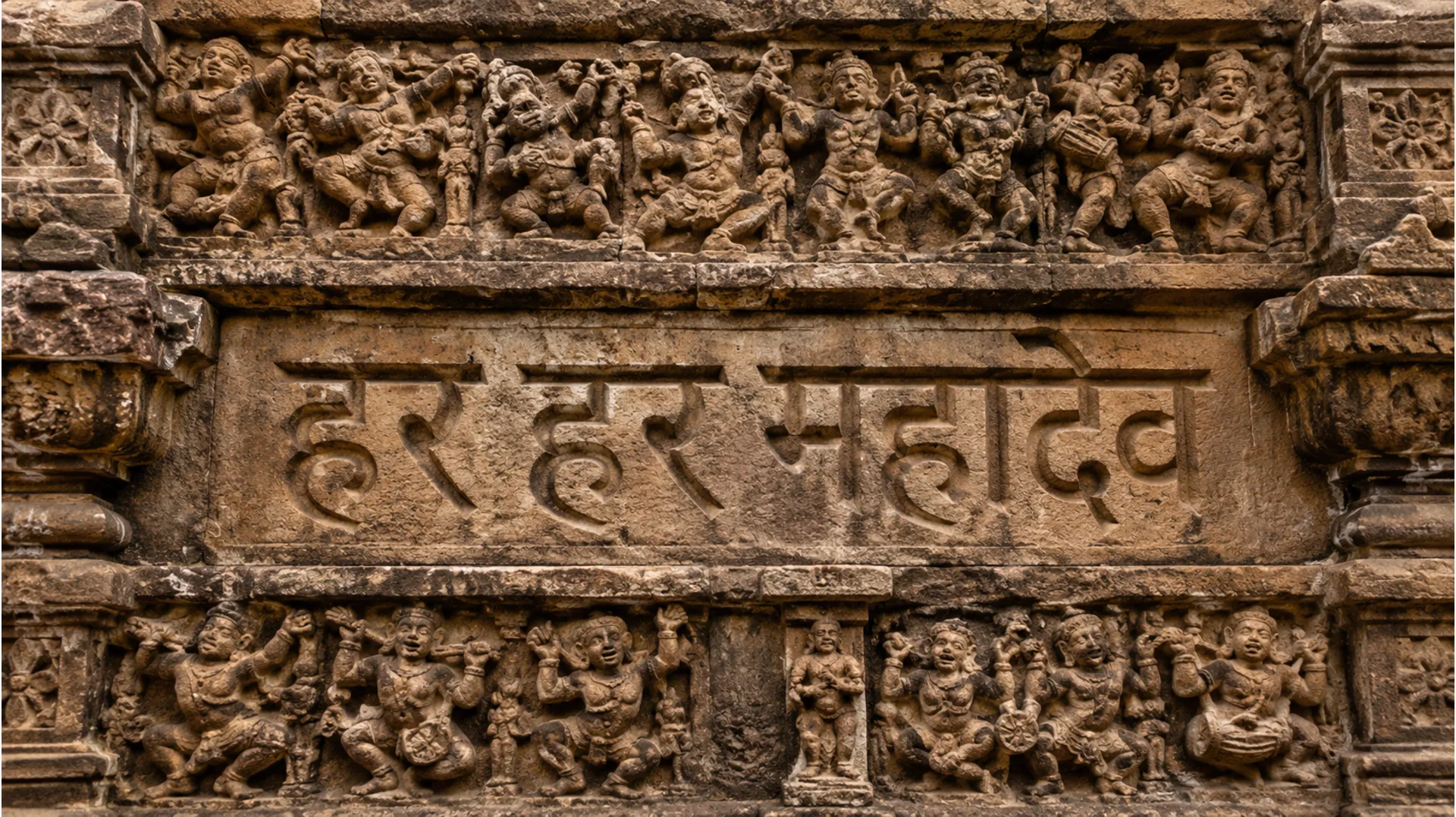 A detailed clear photo camera close-up of worn, weathered sandstone carving on an ancient Indian temple wall, showing the Devanagari script for 'हर हर महादेव' (Har Har Mahadev) surrounded by sculptures of dancing figures.