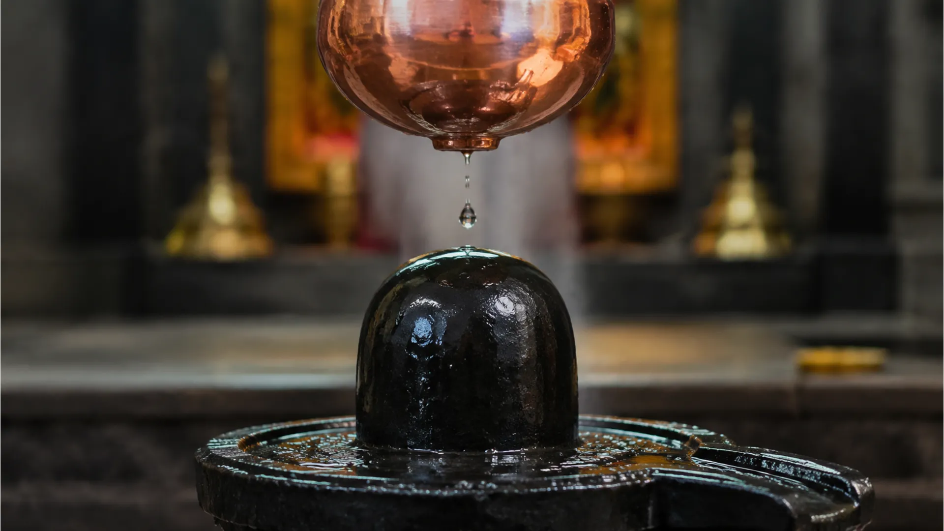 Hanging copper pot dropping water onto a dark stone image shivling